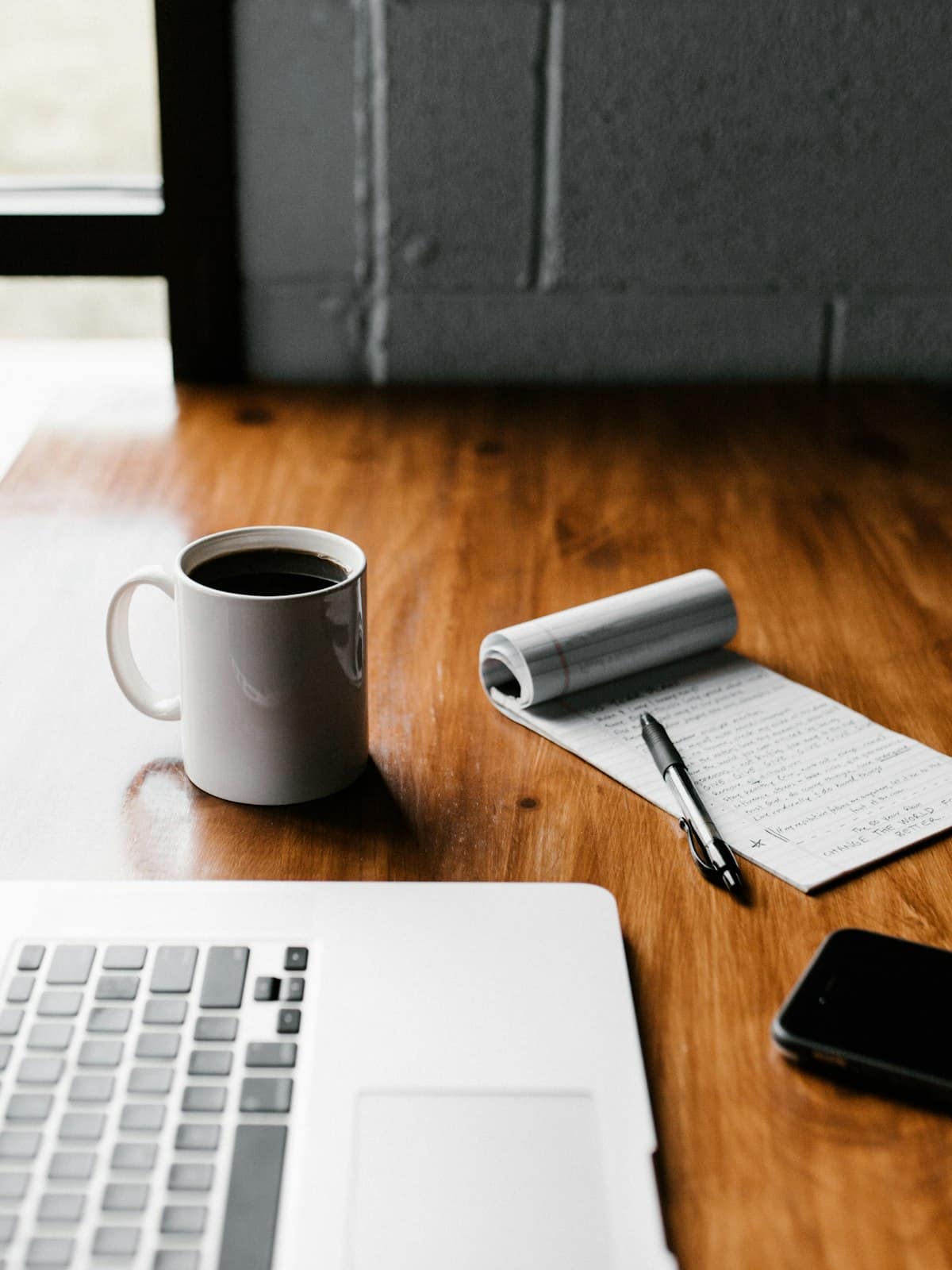 A laptop, coffee, and a notepad on a warm wooden desk by a window.